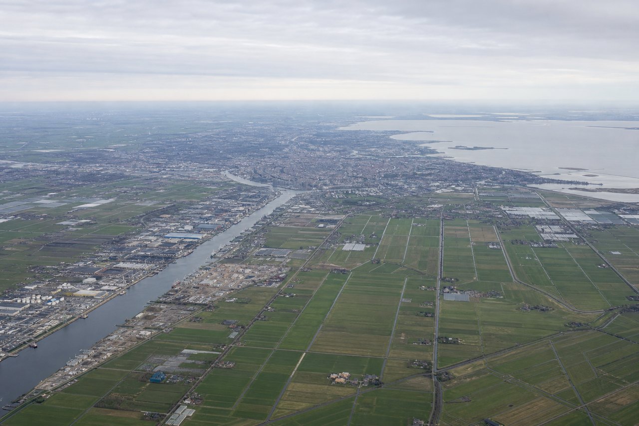 Left window seat view during takeoff from Amsterdam (AMS) — North Sea coastline and dunes, Zandvoort and Noordwijk beach towns, Haarlemmermeer polder greenhouses, IJmuiden harbor entrance further north