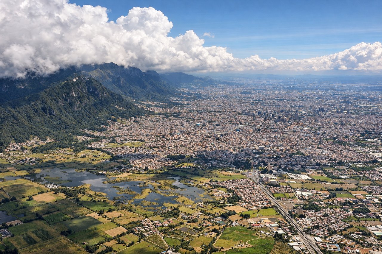 Aerial window seat view during landing at Bogota (left side) — Eastern Cordillera of the Colombian Andes rising steeply to the left and ahead, Bogotá savanna (Altiplano Cundiboyacense) spreading below as a high-altitude plateau at ~2,600 m elevation with patchwork agricultural fields, Monserrate peak (3,152 m) visible as a distinctive forested ridge east of the city, Bogotá's urban sprawl filling the valley floor with a dense grid of buildings and streets