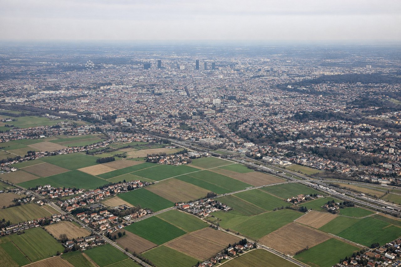 Aerial window seat view during takeoff from Brussels (right side) — Meuse River (visible as a winding ribbon southeast of Brussels), Walloon industrial towns (Charleroi region), early views of the Ardennes plateau edge, Brussels-Zaventem airport infrastructure receding below