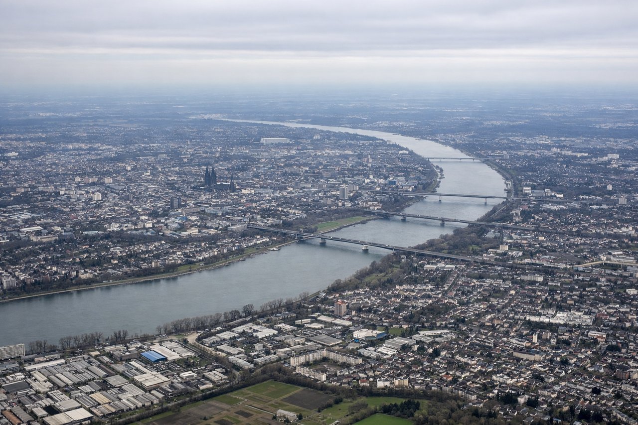 Left window seat view during takeoff from Cologne (CGN) — Cologne Cathedral (Kölner Dom), Rhine River bends through the city, Cologne skyline including Deutz district, Hohenzollern Bridge crossing the Rhine