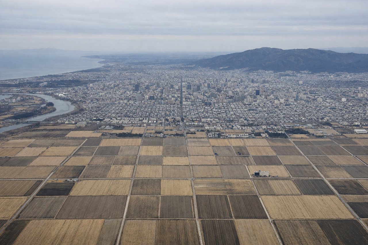 Aerial window seat view during landing at Sapporo (left side) — Lake Shikotsu (caldera lake with distinctive oval shape), Sapporo city grid and urban sprawl, Ishikari Plain agricultural fields, Daisetsu Mountains (distant, to the east)