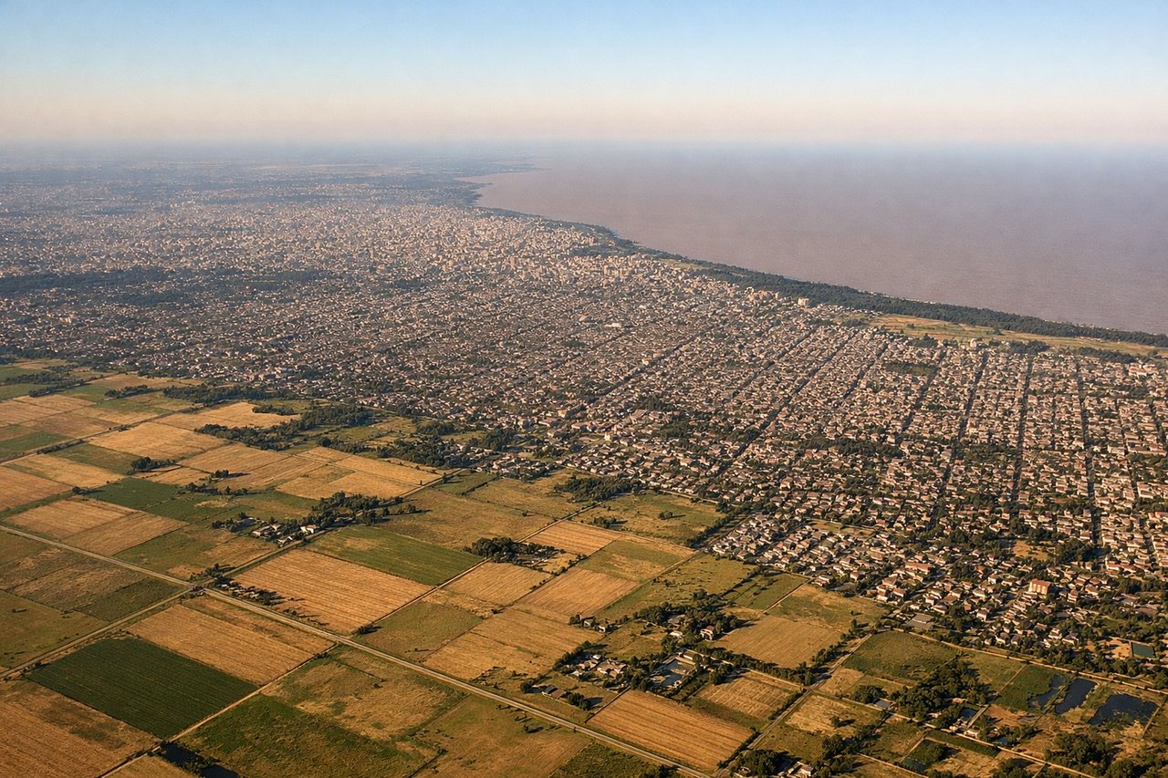 Aerial window seat view during landing at Buenos Aires (right side) — Rio de la Plata estuary (one of the widest rivers in the world, visibly brown from sediment), Buenos Aires urban grid extending to the riverbank, Riachuelo river mouth at La Boca district, flat Pampas grassland stretching west under the aircraft