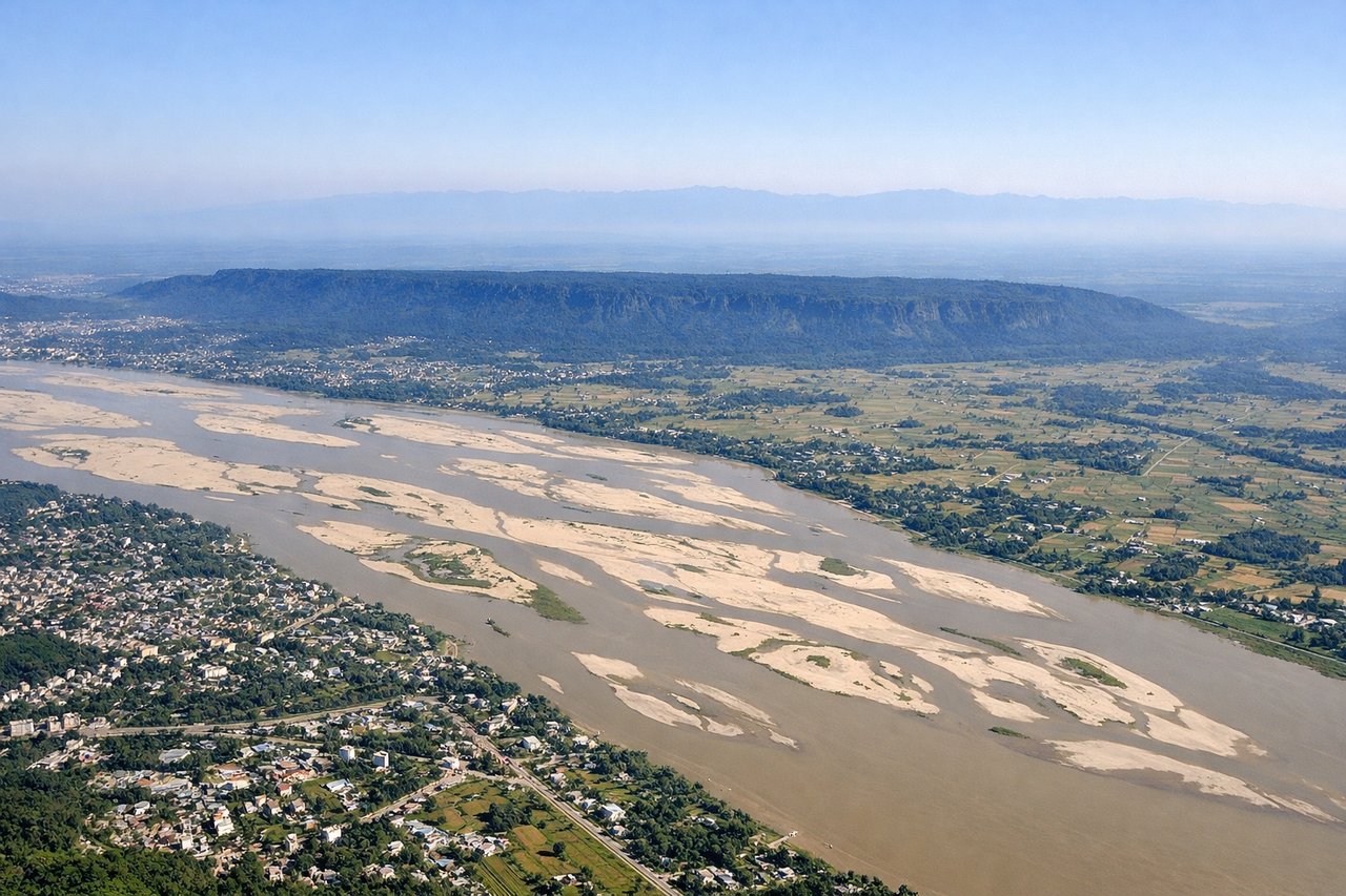 Aerial window seat view during takeoff from Guwahati (left side) — Brahmaputra River with its wide meanders, Guwahati city grid and urban development, Assam plains extending to the horizon, scattered tea gardens and agricultural fields
