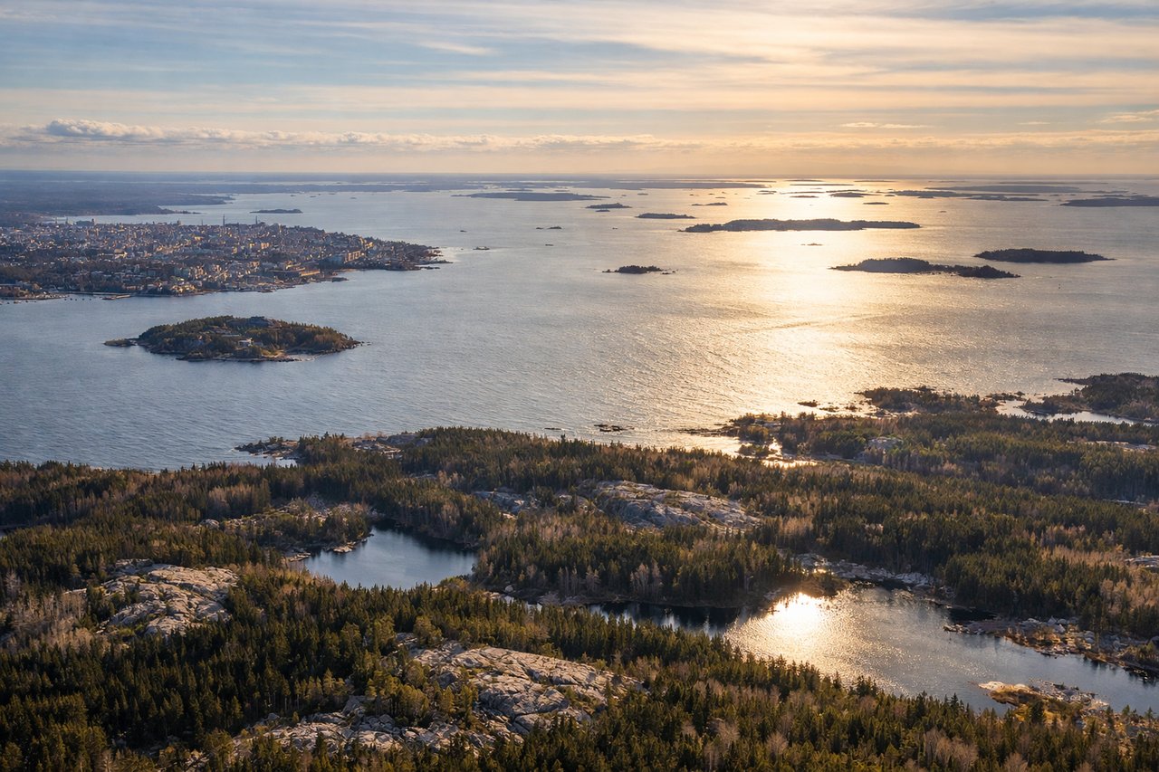 Aerial window seat view during landing at Helsinki (left side) — Baltic Sea and archipelago (islands and skerries visible as dark patches in blue water), Lake Tuusula (large freshwater lake inland, northwest of airport), Finnish boreal forest (dense green coverage across the landscape), Vantaa River valley (threading through forested terrain toward the airport)