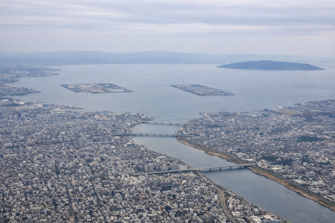 Right window seat view during takeoff from Osaka (ITM) — Osaka Bay (immediate left), Kobe skyline and port facilities (far left on horizon), Rokko Mountains (left horizon), Osaka city grid and Dotonbori district (right and below)
