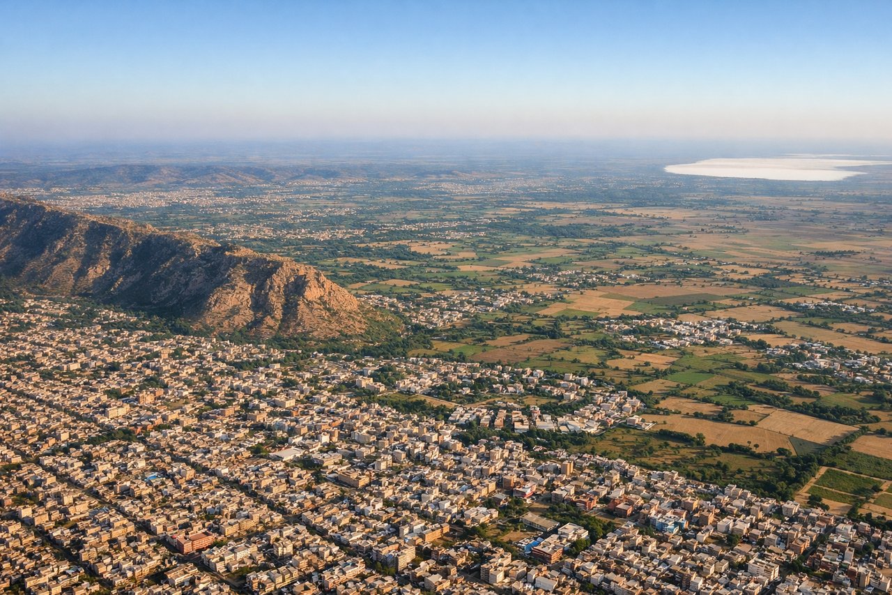 Aerial window seat view during takeoff from Jaipur (right side) — Aravalli Range (foothills), Jaipur city grid and urban sprawl to the left, agricultural plains extending south, Sambhar Salt Lake (seasonal water body visible as white/pale expanse approximately 60 km southwest)