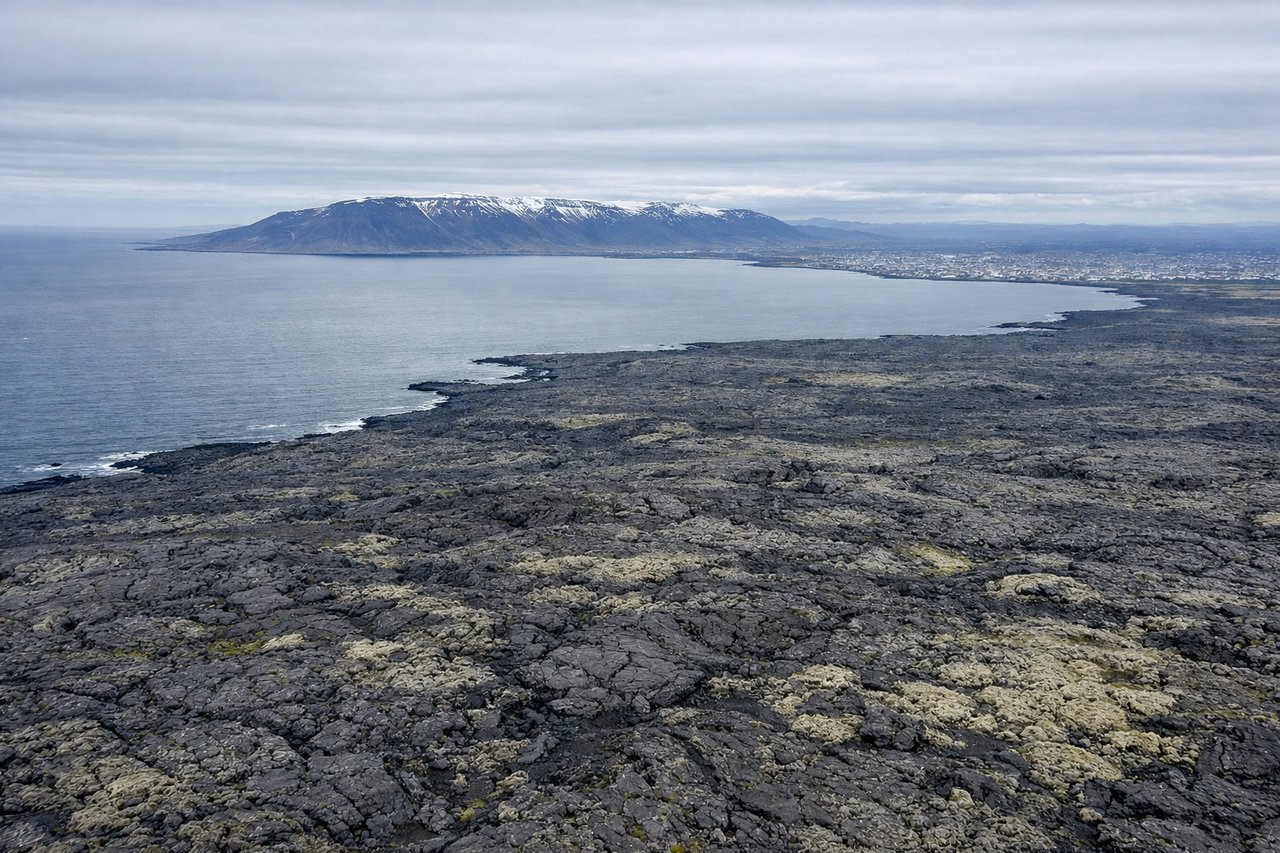 Aerial window seat view during takeoff from Keflavik (left side) — Reykjanes Peninsula lava fields, Faxaflói Bay, Mount Esja across the bay to the northeast, Reykjavik city lights if departing in low light