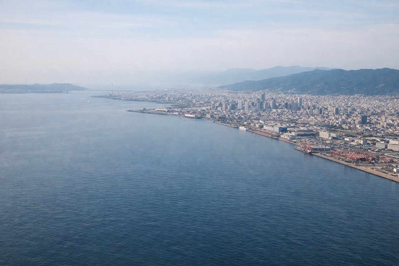 Aerial window seat view during landing at Osaka (right side) — Osaka Bay (dark blue water with shipping traffic), Kansai International Airport's artificial island and parallel runways (distinctive geometric form), Kobe city and port facilities (west across the bay), Awaji Island (south in the distance)