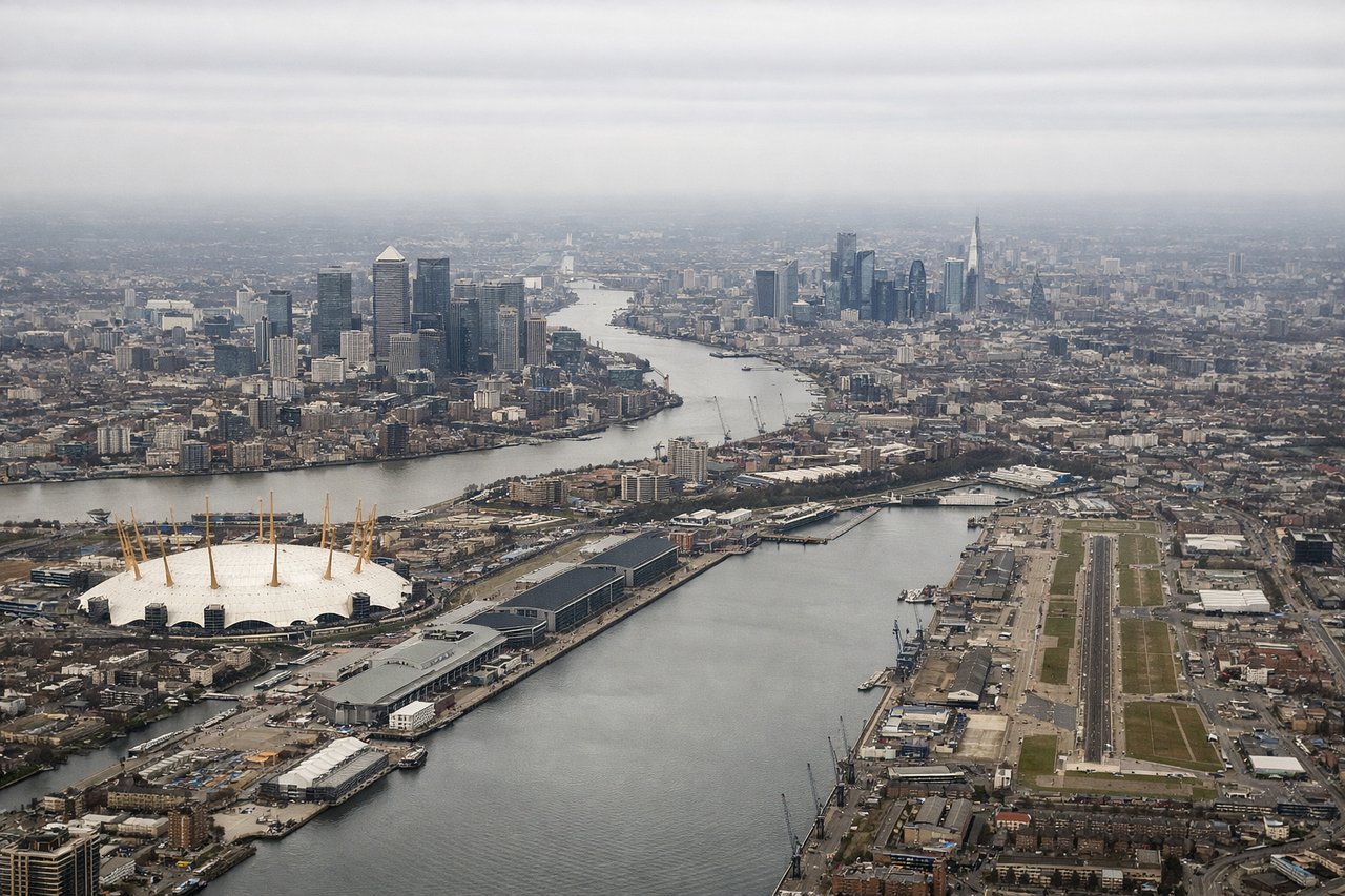 Aerial window seat view during takeoff from London (right side) — River Thames bend at Greenwich, O2 Arena dome, Greenwich Peninsula, Thames Estuary widening toward the North Sea