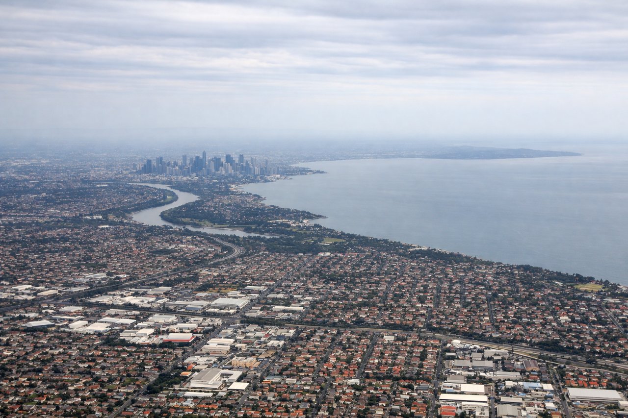 Aerial window seat view during takeoff from Melbourne (right side) — Dandenong Ranges (forested ridge), Port Phillip Bay (water body to the west), Yarra River valley (threading through suburbs), Macedon Ranges (distant peak on horizon to the northwest)
