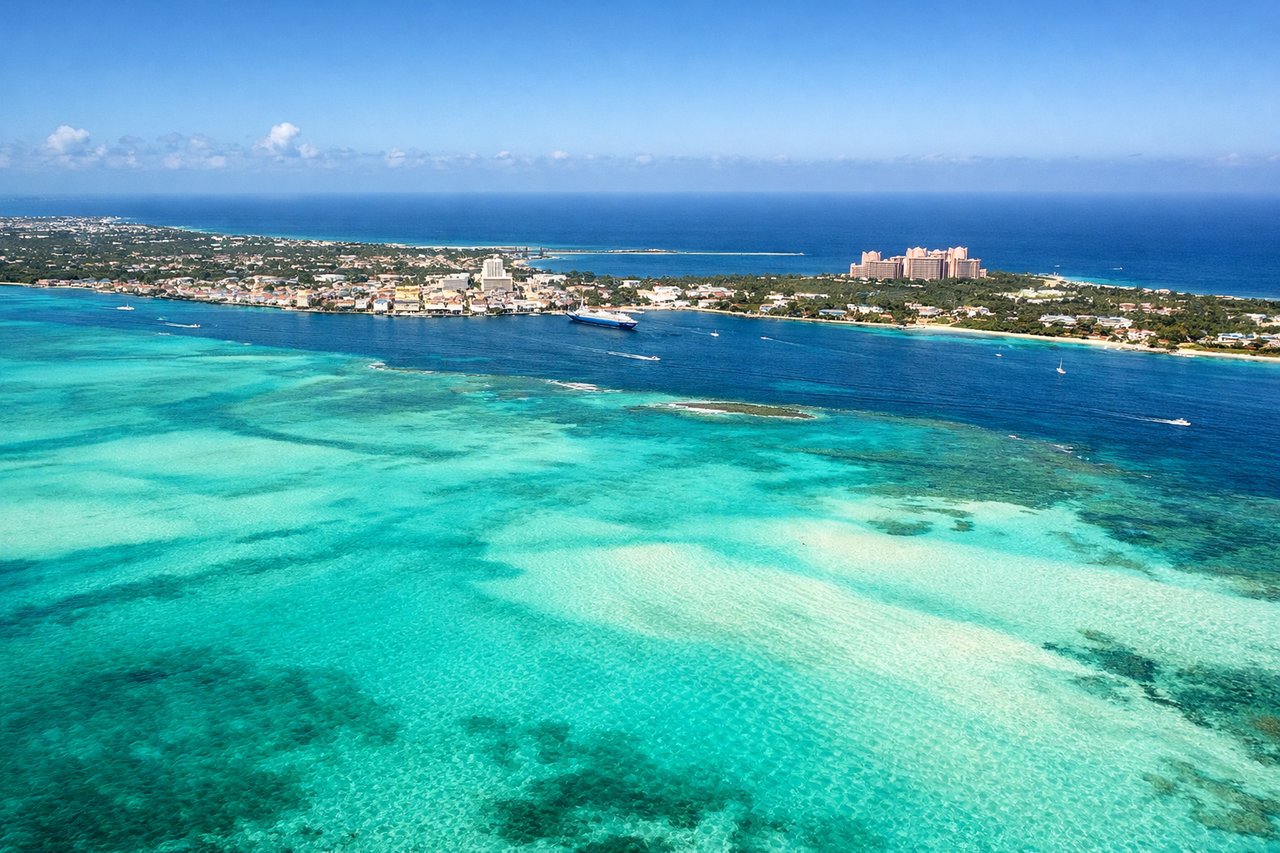 Aerial window seat view during landing at Nassau (left side) — Nassau Harbor (deep blue water with visible boat traffic), Paradise Island (low-lying island with resort development, north of harbor), Bahama Banks (brilliant turquoise shallow water surrounding the islands), New Providence Island (the main island, appearing as low scrubland)