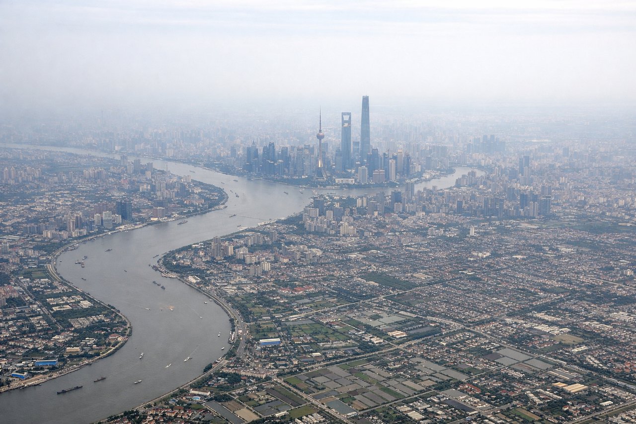 Aerial window seat view during takeoff from Shanghai (right side) — Huangpu River (sharp meander visible below), Pudong financial district towers (Oriental Pearl TV Tower, Shanghai Tower), Hangzhou Bay to the southeast, East China Sea coastline