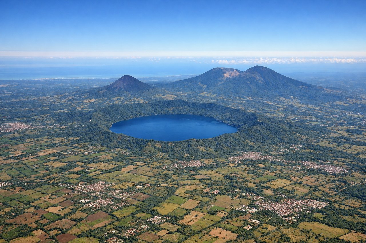 Aerial window seat view during takeoff from San Salvador (right side) — San Salvador volcano (2,422 m, dominant peak southeast of airport), Lake Ilopango (large crater lake visible 15 miles east), Cuscatlán mountain range (extending northeast), Pacific coastal plain (stretching south toward the coast)