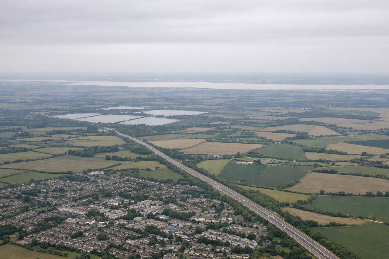 Left window seat view during takeoff from London (STN) — Blackwater Estuary, Essex farmland grid pattern, Clacton-on-Sea coastal strip, North Sea horizon