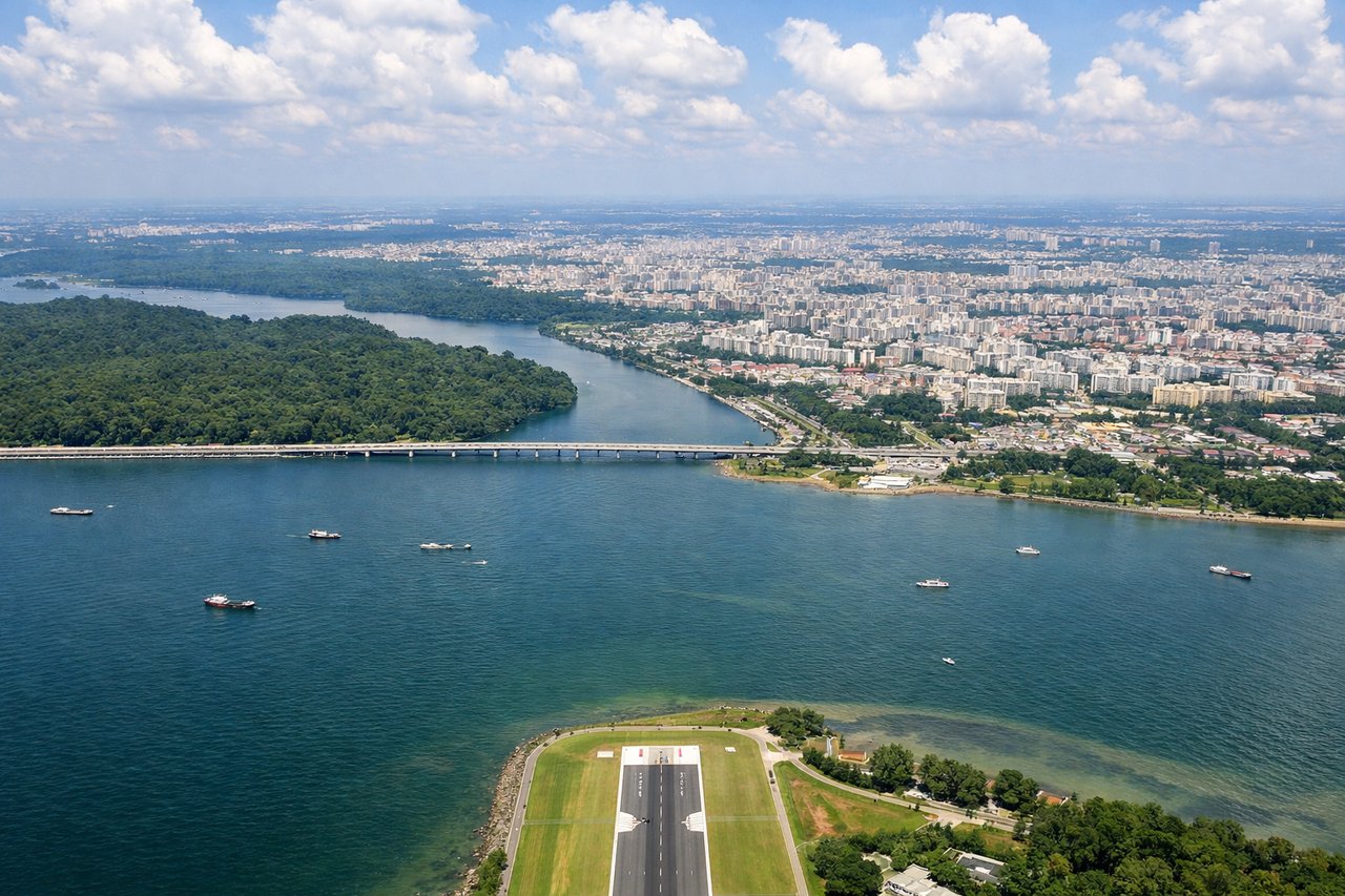 Right window seat view during landing at Singapore (XSP) — Strait of Johor (water body to the right), Johor Bahru cityscape (Malaysian coast across the strait), Seletar Reservoir (inland to the left, visible as a large water body), northern Singapore's residential and industrial zones
